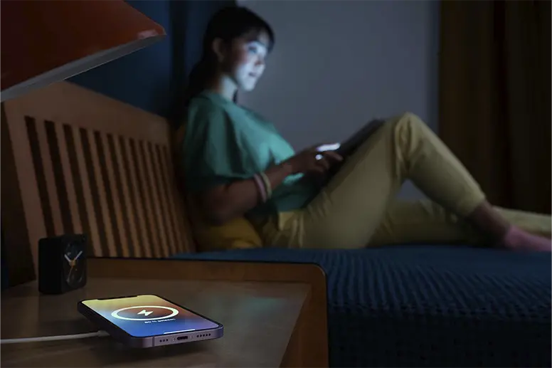 Mobile phone lies on an inductive charging station at night, woman in the background sits on the bed