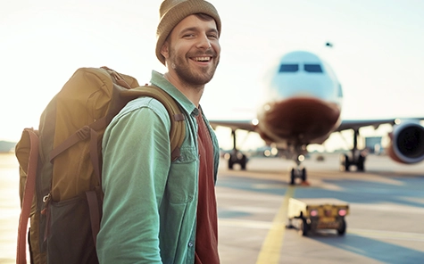 Un bărbat zâmbește la cameră, stând pe un aerodrom cu un avion în fundal. Bărbatul poartă o șapcă tricotată, rucsac, cămașă și tricou.