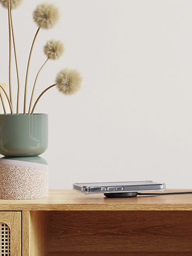 The wireless Qi2 charger with inductive charging pad from Hama lies on a wooden sideboard, a smartphone rests on it and is being charged. Next to it are two vases in pastel colours with dried flowers in them.