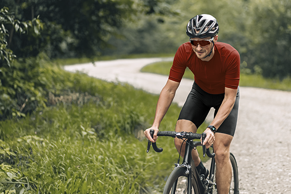 A man in short cycling clothing is sitting on a racing bike and cycling along a gravelled forest path. He is wearing a Hama smartwatch on his wrist.